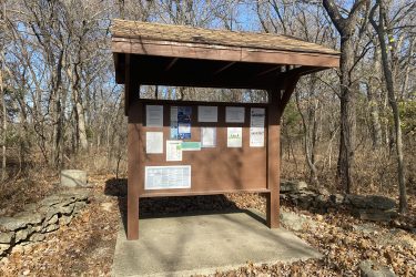 A wooden information kiosk with a sloped roof, situated in a forested area with bare trees and fallen leaves. The kiosk displays multiple printed notices and maps on its sides. Stone structures can be seen at the base of the kiosk, and the ground beneath it is concrete. Drywood Creek Trail mountain bike trail.