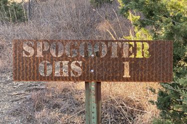 A weathered metal sign displaying the words "SPOGooter OHS 1," mounted on a post in a natural outdoor setting with dry grass and shrubbery in the background. The sign has a rusted finish, adding to its rustic appearance. Fancy Creek State Park mountain bike trail.