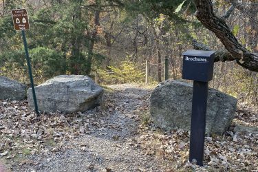 A gravel pathway leading into a wooded area, marked by a sign that indicates the entrance to the National Historic Trails at Alcove Spring. To the right, there is a black brochure box positioned near large rocks, with fallen leaves scattered along the ground. The landscape features trees with autumn foliage in the background. Alcove Springs mountain bike trail.