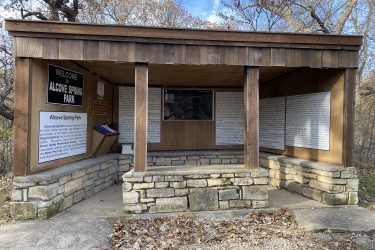 A wooden structure serving as an information kiosk at Alcove Spring Park, featuring a stone foundation. The kiosk has multiple informational panels detailing the park's history and natural features, with a map displayed inside. Surrounding trees and a gravel path are visible. The sky is partly cloudy, indicating a clear day. Alcove Springs mountain bike trail.