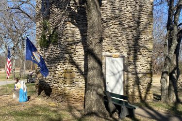 A stone building with a flat roof, surrounded by bare trees, flags, and a bench. One flag is an American flag, and the other is a state flag. A person dressed in historical attire is sitting on the ground nearby, with a grassy area and a path visible in the foreground. Drywood Creek Trail mountain bike trail.