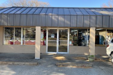 Front view of a retail store with glass doors and large windows displaying various items inside. The signs indicate that the store is open, and the building has a metal roof and beige siding. A few bicycles are visible through the windows, along with other merchandise. There is a small potted plant at the entrance and a vehicle parked nearby.
