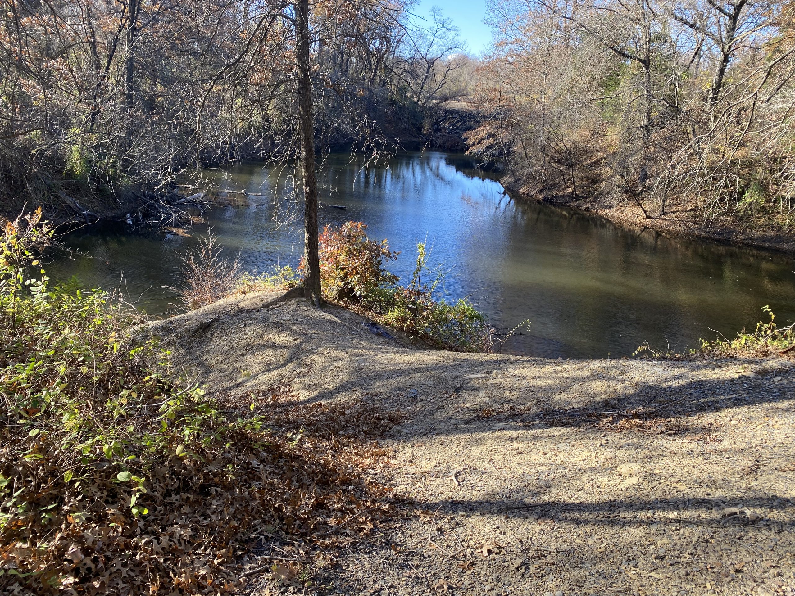 A serene view of a calm river surrounded by bare trees and overgrown vegetation, with a gravelly bank leading down to the water. Sunlight casts shadows on the ground, highlighting the earthy tones of fallen leaves and soil. 23rd St Bike Park mountain bike trail.