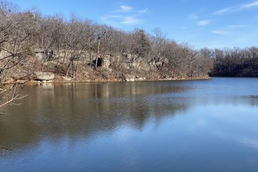 A serene view of a calm lake surrounded by bare trees and rocky cliffs, with a clear blue sky reflecting on the water's surface. The landscape conveys a peaceful, natural setting. Drywood Creek Trail mountain bike trail.