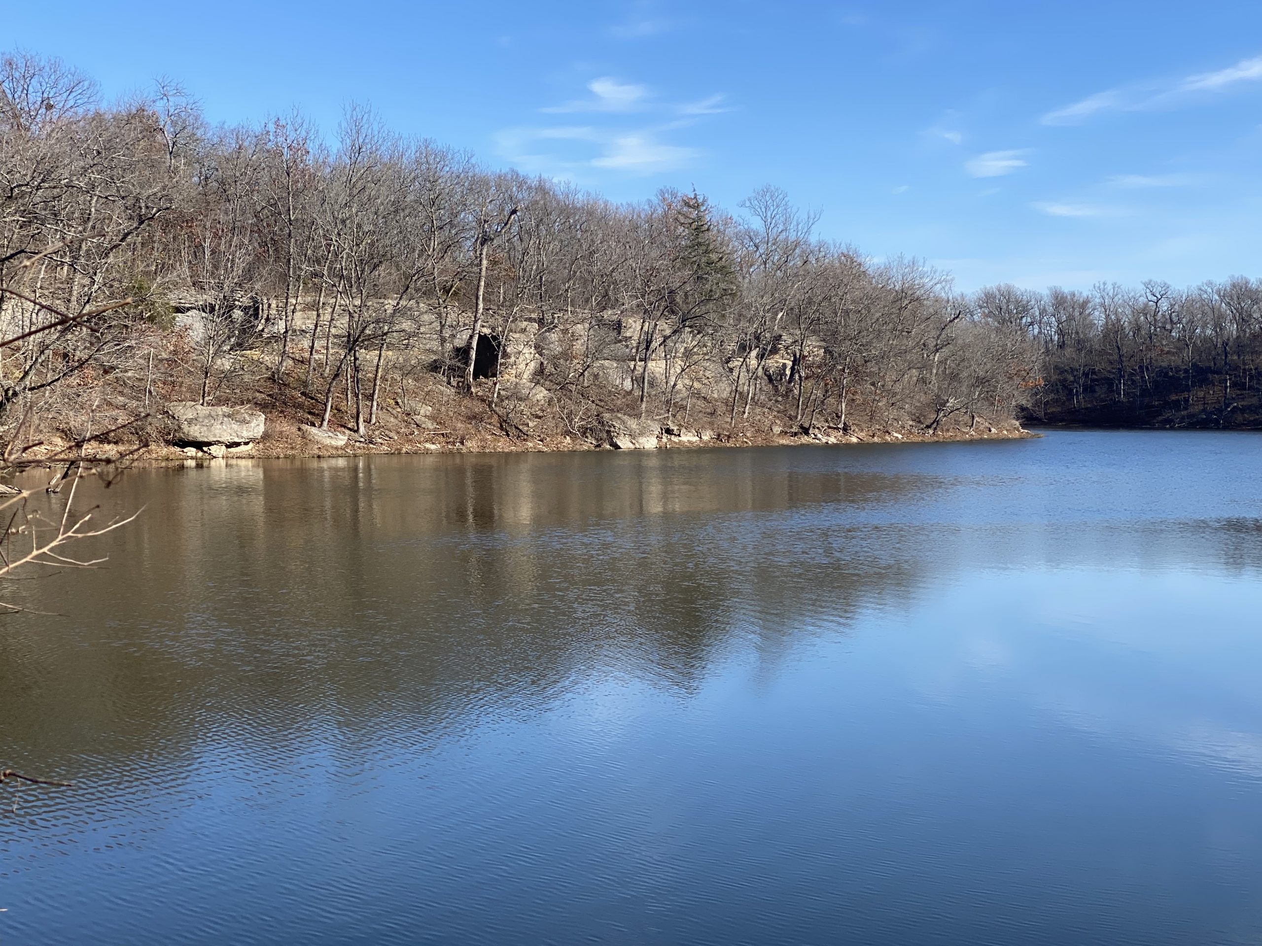 A serene view of a calm lake surrounded by bare trees and rocky cliffs, with a clear blue sky reflecting on the water