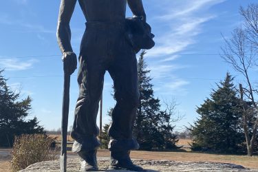 A bronze statue of a farmer or laborer holding a hat and a walking stick, standing on a large rock base. A plaque at the base reads, "C.C.C. Volunteer. We dedicate this monument to the individuals in the Civilian Conservation Corps who contributed to the accomplishments the rest of the future to enjoy. This is the home of Camp Farlington, 1935-1939." The background features a clear blue sky and distant trees. Drywood Creek Trail mountain bike trail.