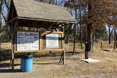 A wooden shelter at the 23rd Street Bike Park displaying trail difficulty levels and park rules. A blue trash can is located in front of the shelter, and a bike pump station is visible nearby. The background features trees and a gravel path. 23rd St Bike Park mountain bike trail.