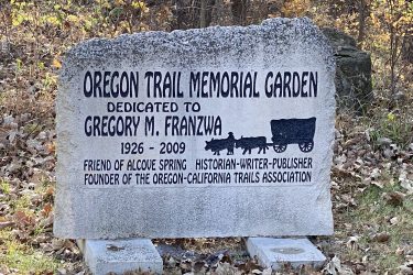 A granite memorial stone commemorating Gregory M. Franzwa, dedicated to the Oregon Trail Memorial Garden. The stone includes engraved text stating his years of life (1926-2009) and highlights his contributions as a historian, writer, publisher, and founder of the Oregon-California Trails Association. Accompanied by a design of a wagon and animals, the stone is set against a backdrop of autumn foliage. Alcove Springs mountain bike trail.