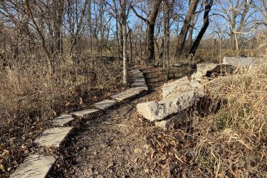 A winding stone path surrounded by bare trees and dry grass in a natural setting on a clear day. Cottonwood River Trail mountain bike trail.