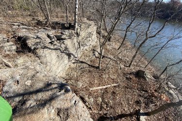 A rocky area with scattered leafless trees, overlooking a calm body of water. The terrain is uneven and features various sizes of rocks, with some exposed roots and dried leaves on the ground, indicating a natural landscape in late autumn or winter. Drywood Creek Trail mountain bike trail.
