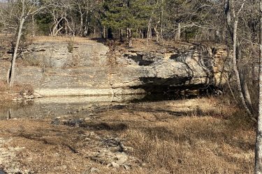 Rocky terrain with a cliff face partially covered by trees, reflecting in a still body of water, surrounded by dry grass and scattered rocks. Drywood Creek Trail mountain bike trail.