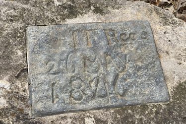 A weathered metal plaque embedded in a rock, inscribed with the text "J.T. Reed 20 M.A. 1846." The surrounding area features scattered dried leaves and a rocky surface. Alcove Springs mountain bike trail.