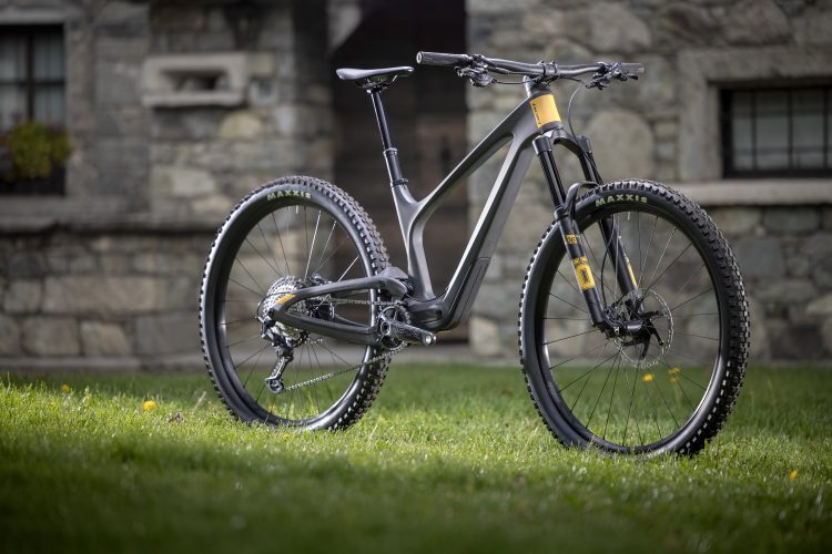 A modern mountain bike displayed on green grass with a stone building in the background. The bike features a sleek black frame with yellow accents, prominent Maxxis tires, and a streamlined design ideal for off-road cycling.