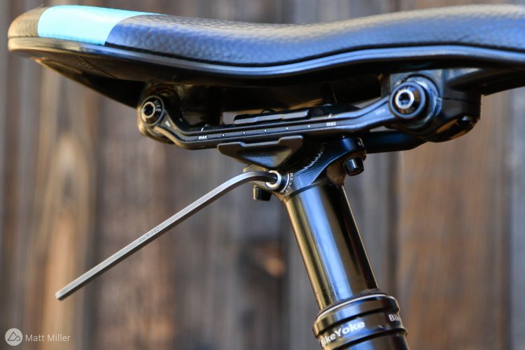 Close-up of a bicycle seat post and saddle, highlighting the adjustment mechanism with a tool inserted. The saddle features a sleek design with a textured surface, and there are visible markings for adjustment limits on the mechanism. The background is blurred wooden texture.