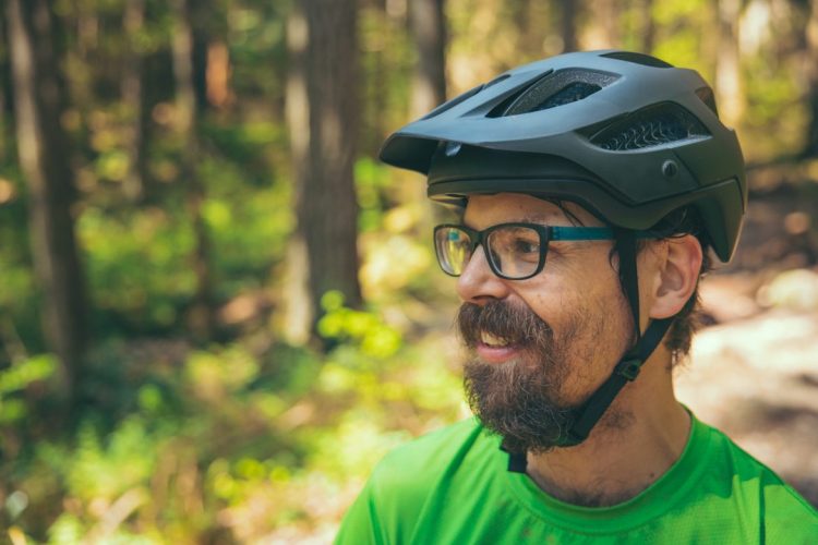 A smiling man wearing a black cycling helmet and glasses stands outdoors, surrounded by greenery in a forest setting. He is dressed in a bright green shirt, and the warm sunlight filters through the trees behind him.