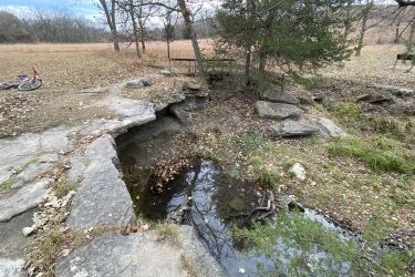 A rocky edge beside a small, clear pool of water surrounded by fallen leaves and sparse vegetation, with a bicycle resting on the ground nearby. In the background, trees and an open grassy area can be seen under a cloudy sky. Alcove Springs mountain bike trail.