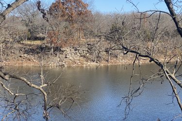 A calm body of water reflecting the clear blue sky, surrounded by a rocky shoreline and partially wooded area with leafless trees. The scenery suggests a tranquil outdoor location, likely during the winter season. Drywood Creek Trail mountain bike trail.