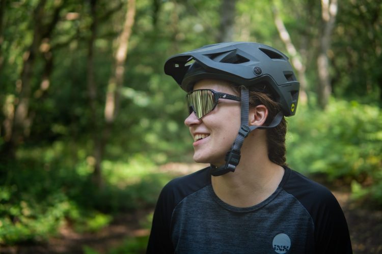 A person wearing a black cycling helmet and reflective sunglasses smiles while standing in a lush green forest. They are dressed in a dark athletic shirt, surrounded by trees and foliage, suggesting an outdoor biking or adventure activity.
