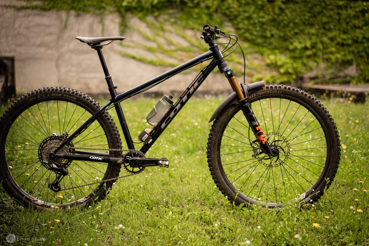 A black mountain bike with thick tires is parked on a grassy area. The bike features a prominent frame design and a water bottle holder attached to the frame. In the background, lush greenery and a textured wall are visible, contributing to a natural, outdoor setting.