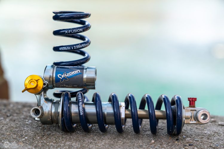 A close-up image of an X-Fusion bike shock absorber, featuring a coiled spring and an adjustable reservoir. The shock is designed for high-performance applications, with clear branding and specifications marked on the spring, indicating a 450 lb/in rate. The background is blurred, suggesting an outdoor setting.