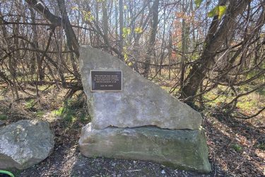A large rock with a bronze plaque commemorating a land donation, surrounded by trees and foliage. The plaque reads, "This land has been donated by Lois and Julian Granato for the preservation of wildlife and the enjoyment of all. June 30, 2000." Wilderness Park Trails mountain bike trail.