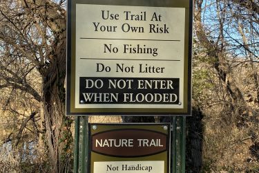 Signage for the Cottonwood River Trail and Nature Trail, featuring warnings to use the trail at your own risk, prohibitions against fishing and littering, and a caution to avoid the area when flooded. The Nature Trail sign also notes that it is not handicap accessible and has steep embankments. The surrounding area includes trees and natural vegetation. Cottonwood River Trail mountain bike trail.
