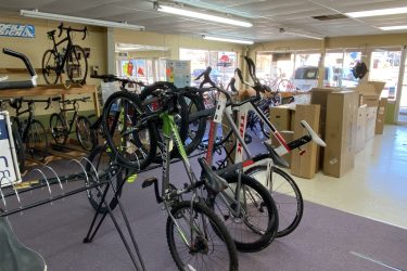 A view inside a bicycle shop showcasing various bicycles on display. Several bikes are positioned on racks, and a few are leaning against each other. Cardboard boxes are visible in the background, indicating new inventory. The shop has a bright, open layout with large windows providing natural light.