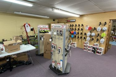 A retail space featuring cycling gear and accessories. On display are racks holding various helmets in different colors, a hydration and gloves display stand, and a table cluttered with boxed items. A cycling jersey hangs on the wall, with additional space for bike maintenance equipment visible. The floor is carpeted, and the lighting is bright, creating an inviting atmosphere for customers.