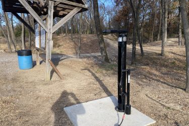 A hand pump stands on a concrete base, surrounded by dirt and trees. In the background, there is a wooden shelter with a sign and a blue trash bin nearby. The scene is set in a park or trail area, with a pathway visible leading into the wooded surroundings. 23rd St Bike Park mountain bike trail.