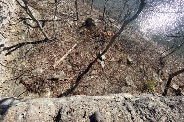 Aerial view of a rocky ledge with a person's boot in the foreground, overlooking a wooded area with bare trees and rocky ground leading down to a reflective body of water. Drywood Creek Trail mountain bike trail.
