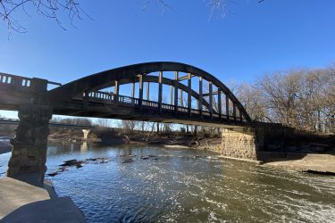 A steel arched bridge spans a river, with clear blue skies overhead. The bridge features stone supports and a wooden deck. The water below reflects sunlight, creating a serene scene with trees lining the riverbank. Cottonwood River Trail mountain bike trail.