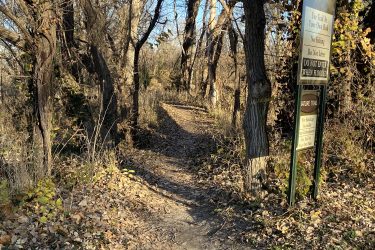 A narrow dirt path winding through trees and shrubs, leading into a natural area. A sign on the right provides trail usage information and warnings, including "Use trail at your own risk" and "Do not enter when flooded." The ground is covered with fallen leaves, indicating an autumn setting. Cottonwood River Trail mountain bike trail.