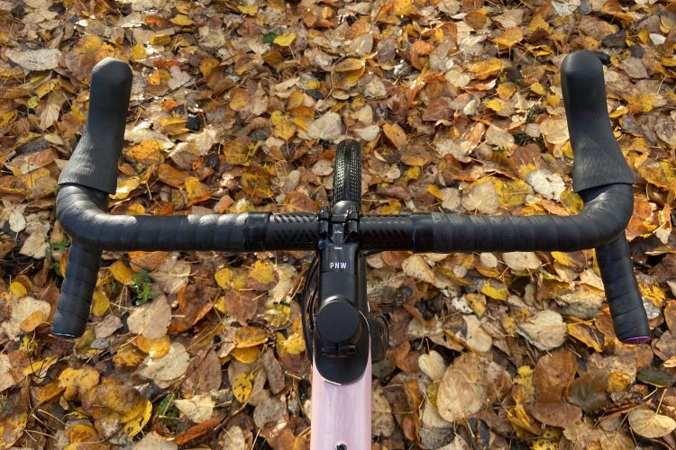 A close-up view of a bicycle handlebar set against a ground covered in autumn leaves. The handlebars are black with textured grips, and the frame of the bicycle is a light pink color. The scene captures the essence of a fall day, highlighting the contrast between the bike and the colorful leaves.