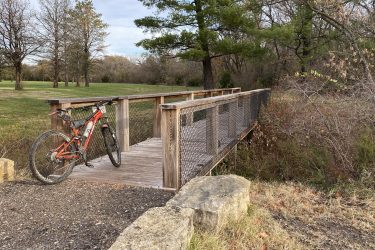 A mountain bike is parked on a wooden bridge surrounded by trees and grassy areas. The scene depicts a peaceful outdoor setting with a clear sky. ESU Trail mountain bike trail.