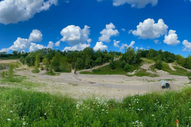 A panoramic view of a lush green area with a sandy landscape under a bright blue sky dotted with fluffy white clouds. The foreground features tall grass and wildflowers, while gentle hills and trees frame the background, creating a serene natural setting. Quarry Trails Metro Park mountain bike trail.