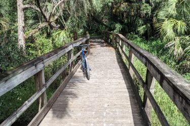 A wooden walking path bridges through a lush green area, surrounded by dense vegetation and trees. A blue bicycle is propped against the railing on the left side of the path, inviting exploration of the natural surroundings. Live Long and Prosper mountain bike trail.
