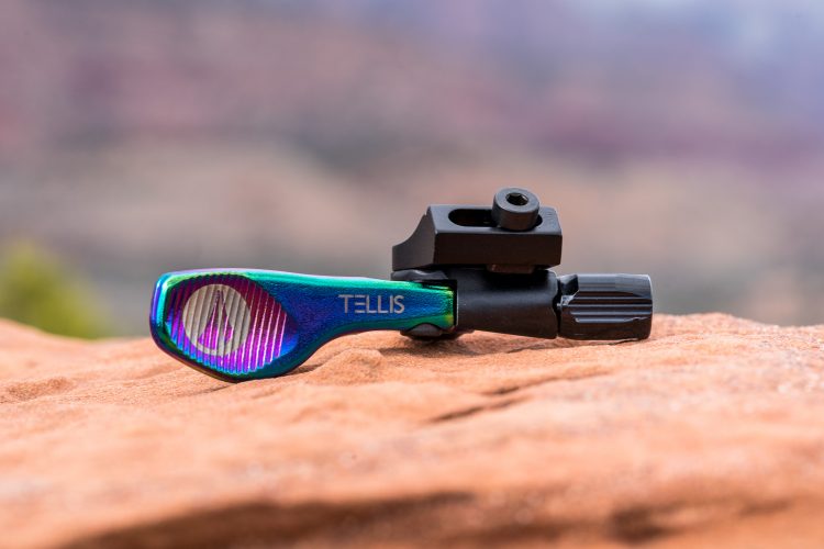 A close-up image of a colorful mechanical device with a blue and green finish resting on a rocky surface. The device features a textured grip and a black attachment component. The background is softly blurred, showcasing a natural outdoor setting.