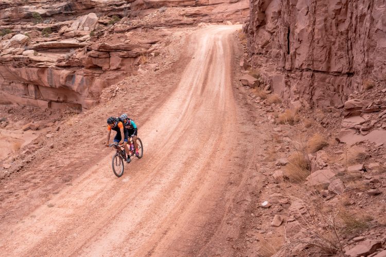 Two mountain bikers ride closely together on a dirt trail surrounded by rocky terrain. The path winds through a desert landscape, with reddish-brown rocks and sparse vegetation. The bikers are wearing helmets and cycling gear, demonstrating an adventurous spirit in an outdoor setting.
