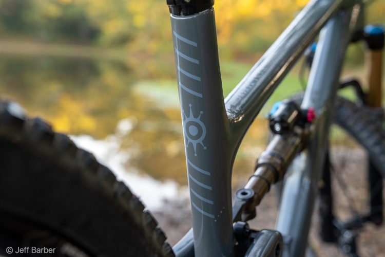 Close-up of a mountain bike frame showcasing the grey finish and a decorative graphic, with a blurred background of a serene pond and autumn foliage. The bike's rear tire is partially visible on the left side, emphasizing its rugged design.
