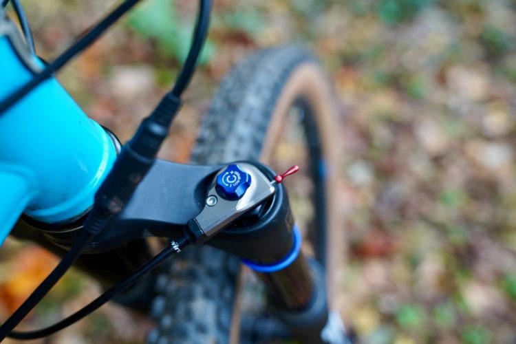 Close-up of a mountain bike's suspension fork with a blue adjustment knob and connected cables, set against a blurred background of autumn leaves.