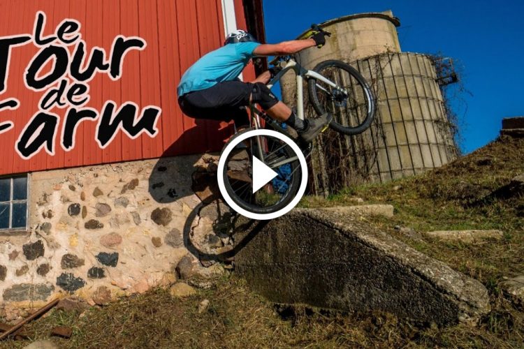 A mountain biker in a blue shirt performs a jump off a stone structure beside a rustic red barn, with a clear blue sky in the background. The scene features the logo "Le Tour de Farm" prominently displayed on the left.