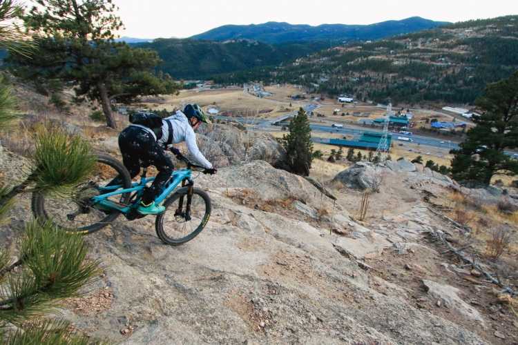 A mountain biker navigating a rocky trail on a hillside, with a panoramic view of a valley and mountains in the background. The cyclist is wearing a helmet and riding a blue mountain bike, while trees and boulders are visible in the foreground.