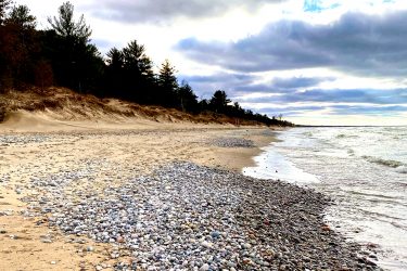 A scenic beach view featuring a stretch of shoreline adorned with smooth, multicolored pebbles. The sandy beach is bordered by patches of trees in the background under a moody sky filled with clouds, suggesting an overcast day. Gentle waves lap at the shore, creating a tranquil atmosphere. Pinery Provincial Park mountain bike trail.
