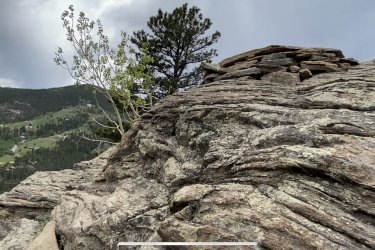 A close-up view of a rocky hillside with layered granite formations, featuring a small pile of stones on top. A green tree is visible in the background, along with a lush landscape of rolling hills and scattered houses under a cloudy sky. The Sluice mountain bike trail.