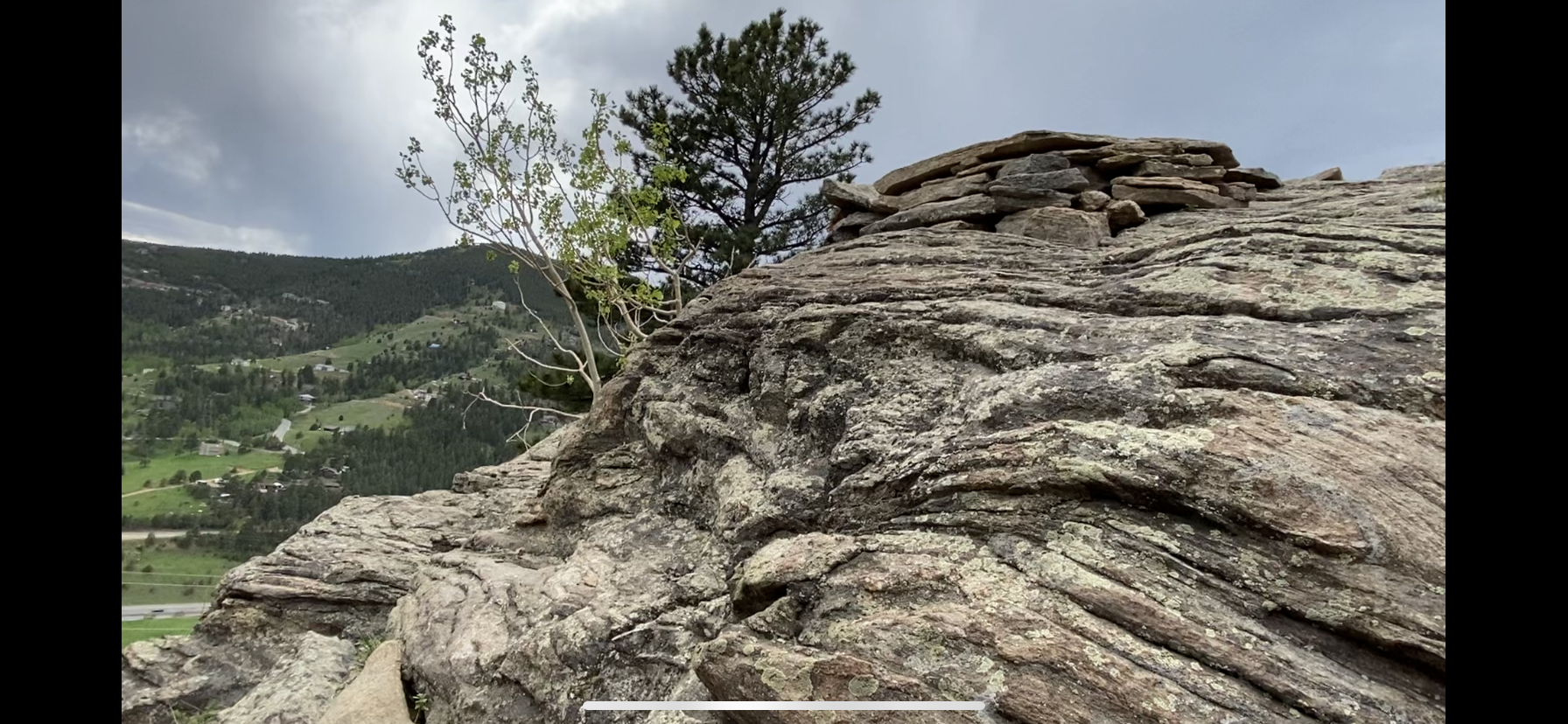 A close-up view of a rocky hillside with layered granite formations, featuring a small pile of stones on top. A green tree is visible in the background, along with a lush landscape of rolling hills and scattered houses under a cloudy sky. The Sluice mountain bike trail.