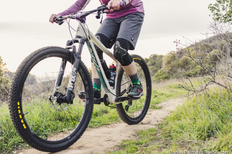 A person riding a mountain bike along a dirt trail, wearing a purple shirt, black shorts, and knee guards. The bike features a prominent front suspension and Maxxis tires. The background includes lush green grass and shrubs, with a soft-focus natural landscape at sunset.