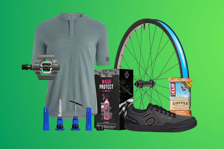 An assortment of cycling-related items displayed on a green background, including a gray short-sleeve jersey, a bike pedal, a bicycle wheel with a blue rim, a Muc-Off Wash Protect and Lube Kit, a set of bike repair tools, a pair of black riding shoes, and a Clif Bar Coffee Collection snack.