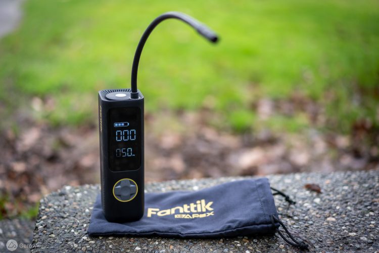 A digital air pump with a display screen showing pressure readings, placed on a textured surface beside a black drawstring bag with the logo "Fanttik" visible. The background features blurred green grass.
