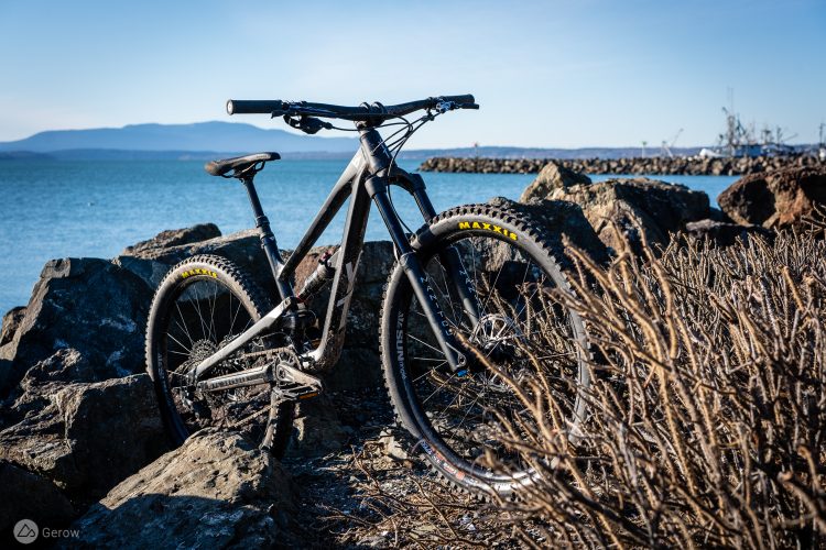 A mountain bike leaning against rocks by a coastal landscape, with calm blue water in the background and mountains in the distance under a clear sky.