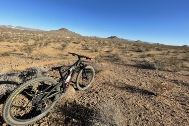 A mountain bike resting on a rocky trail in a desert landscape, with sparse vegetation and distant mountains under a clear blue sky. Tao to Bell Mountain Loop mountain bike trail.